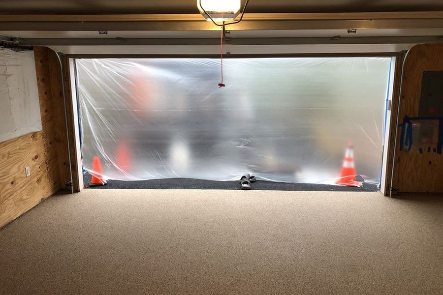 Garage interior with a newly coated speckled tan floor, plastic sheeting and orange cones sealing off the open garage door under an overhead light.