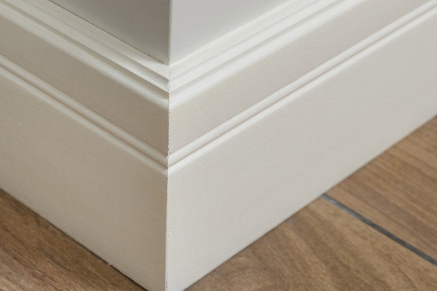 Close-up of a crisp white painted baseboard with layered molding at an outside corner, meeting a warm wood-plank floor.