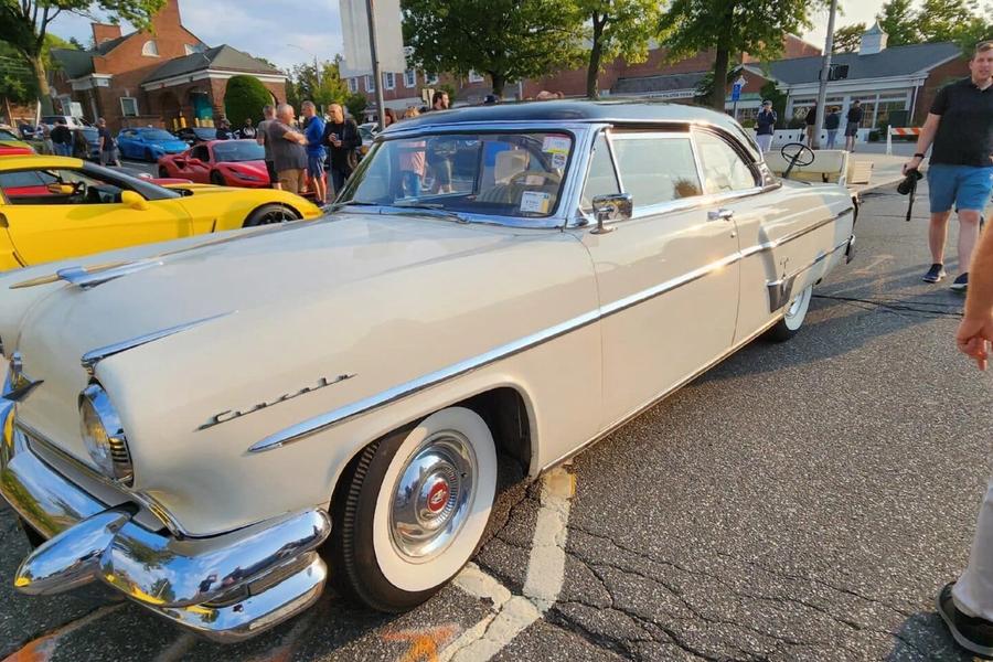 A cream 1950s Lincoln Capri with whitewall tires parked at an outdoor car show, with spectators and a yellow sports car nearby.
