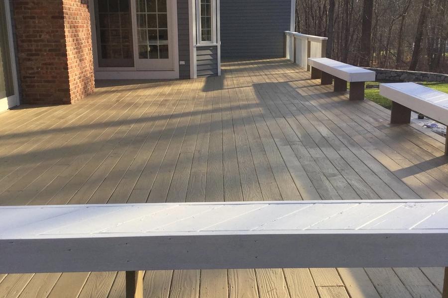 Spacious backyard wood deck with freshly stained tan planks and white-topped built-in benches, attached to a gray-sided house with brick chimney and bare trees behind.