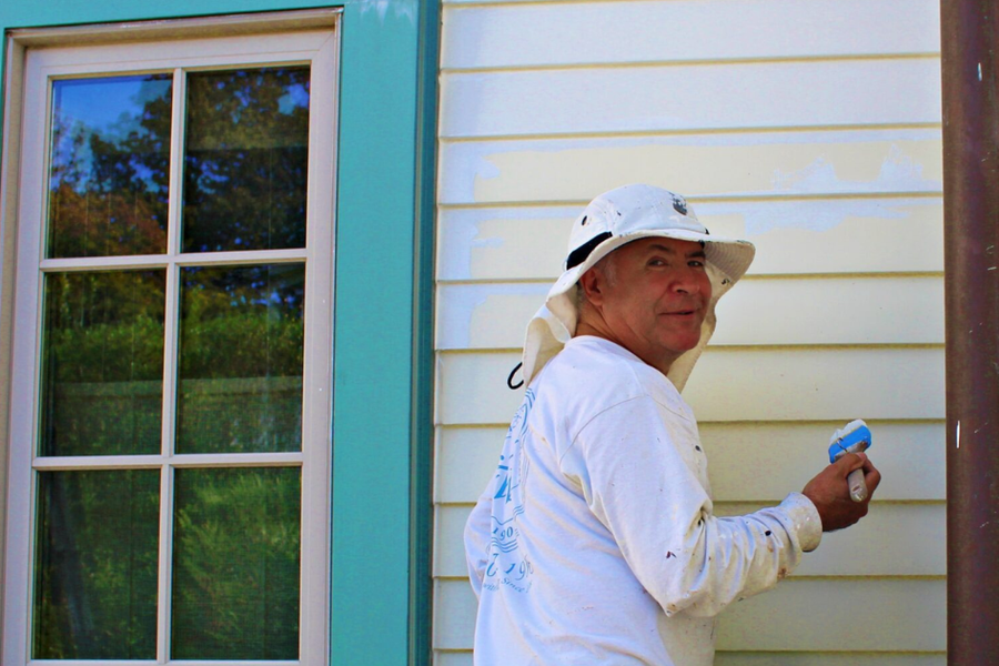 A painter in a white shirt and sun hat applies primer to cream-colored clapboard siding beside a window with teal trim.