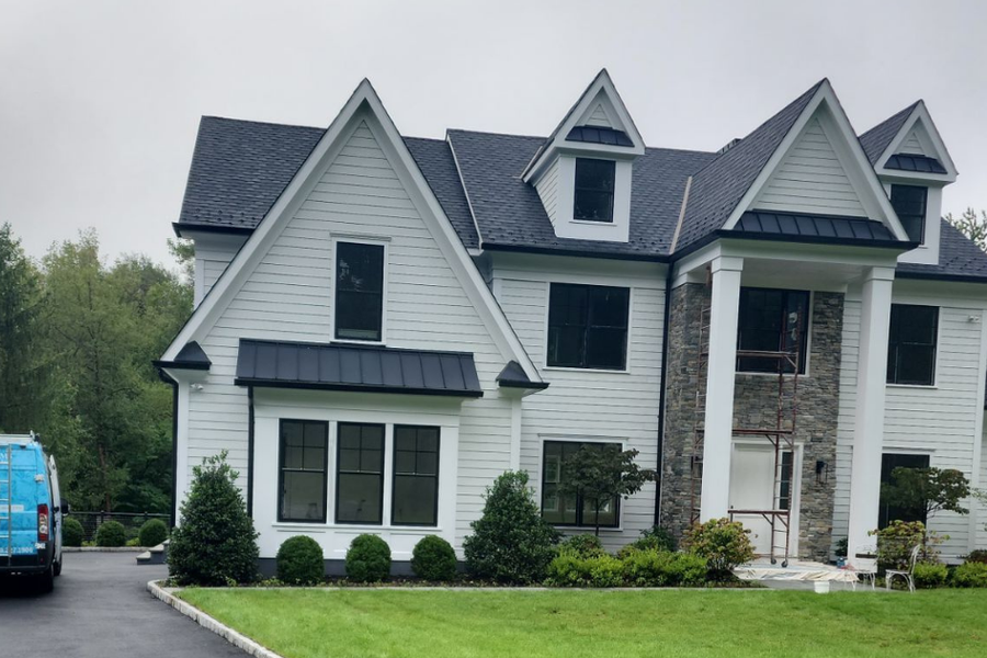 A blue A.G. Williams van parked in the driveway of a large white shingle-style home with black trim, stone accents, and a portico under construction scaffolding.