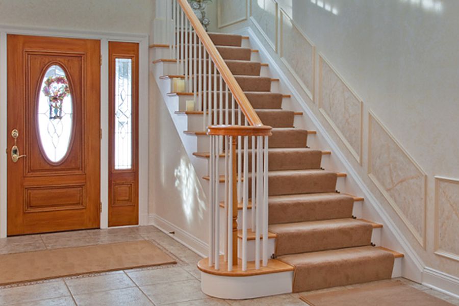 Foyer with a carpeted staircase and white balusters, cream wainscoted walls, tile flooring, and a wood front door with an oval glass panel and wreath.