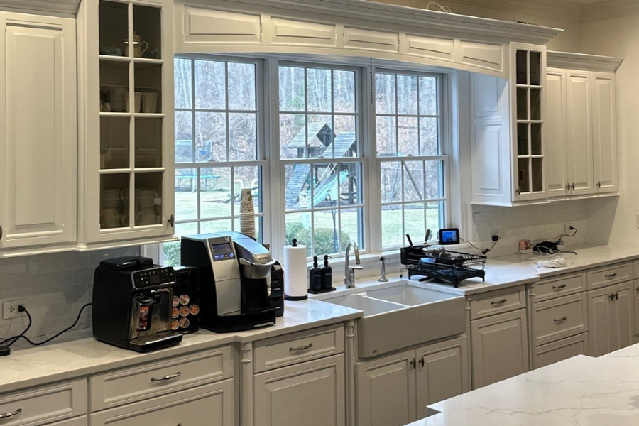Bright kitchen with white painted cabinets, glass-front uppers, marble-look countertops, a farmhouse sink beneath large windows, coffee machines on the counter, and roses on the island.