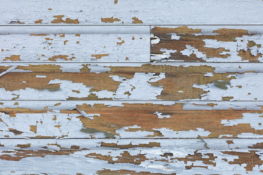 Weathered wood siding with pale blue-gray paint peeling and flaking away, exposing bare brown boards across the surface.