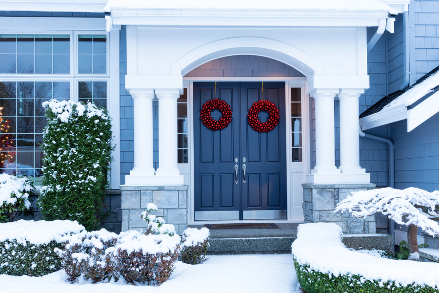 Snow-covered front entrance of a blue-gray house with white columns, an arched portico, and navy double doors decorated with two red berry wreaths.