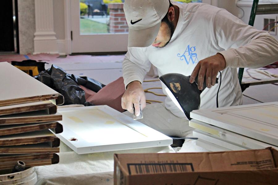 A painter in a white AGW shirt and cap uses a power sander on a white cabinet door laid flat on a protected work surface indoors.