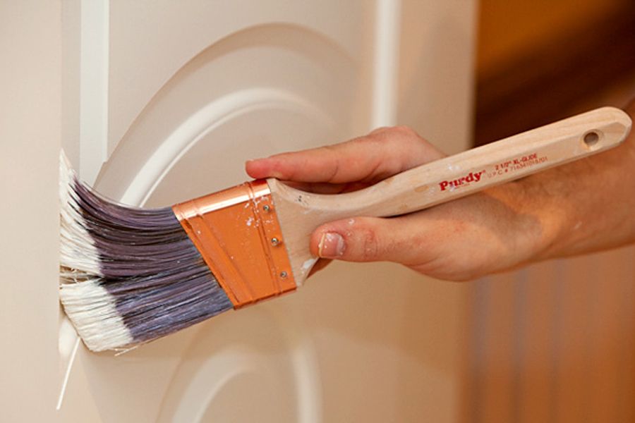 A hand guides an angled Purdy paint brush along the curved molding of a white interior door.