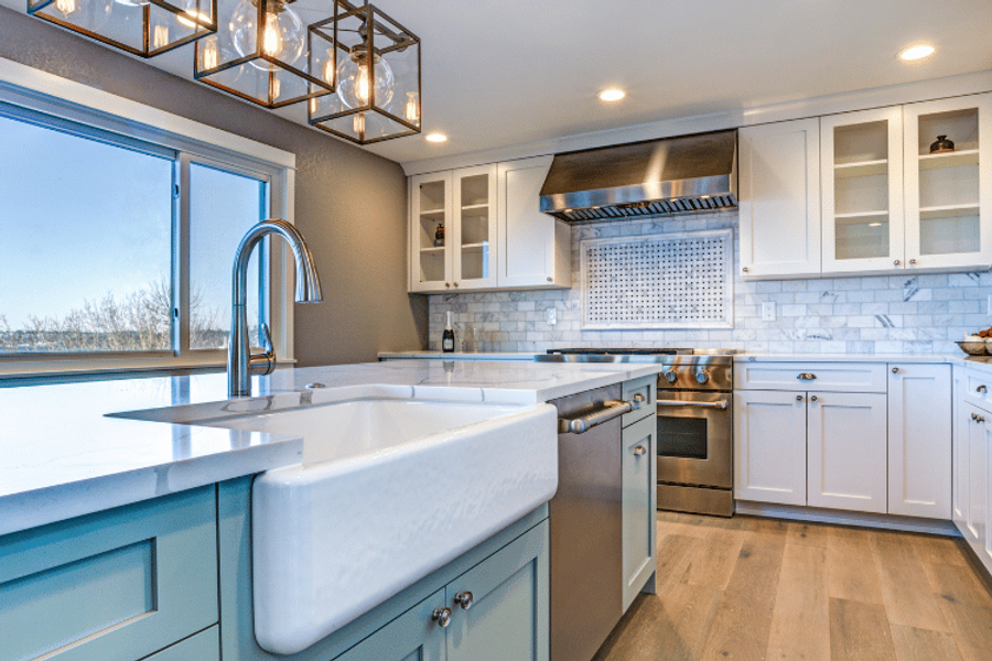 Bright kitchen with white shaker cabinets, light blue island cabinetry, a farmhouse sink, marble tile backsplash, stainless steel range hood, and caged pendant lights over the island.