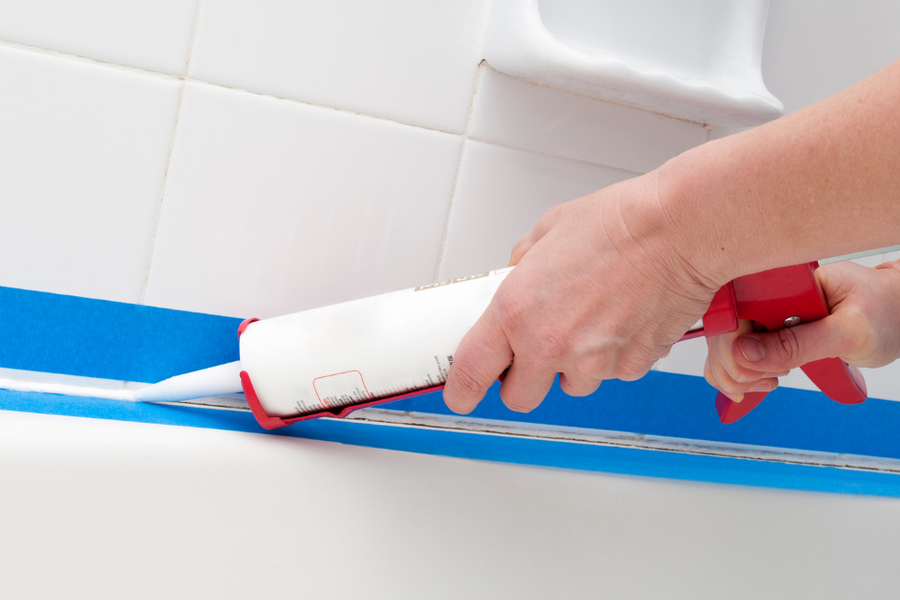 Hands using a red caulking gun to apply a white bead of caulk along a bathtub edge masked with blue painter's tape against white tile.