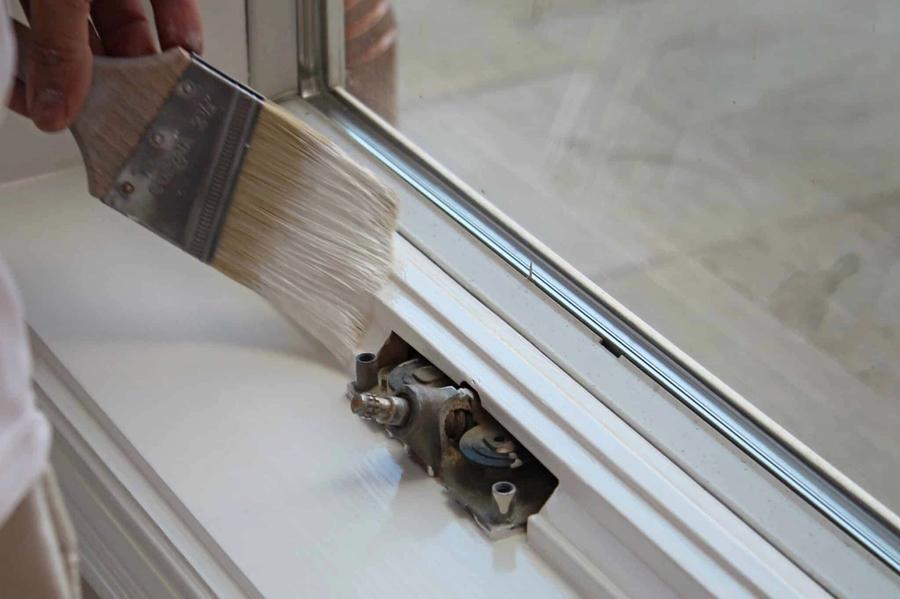 A hand brushes white paint onto a window sill and frame next to the sash lock hardware, with the glass pane visible above.