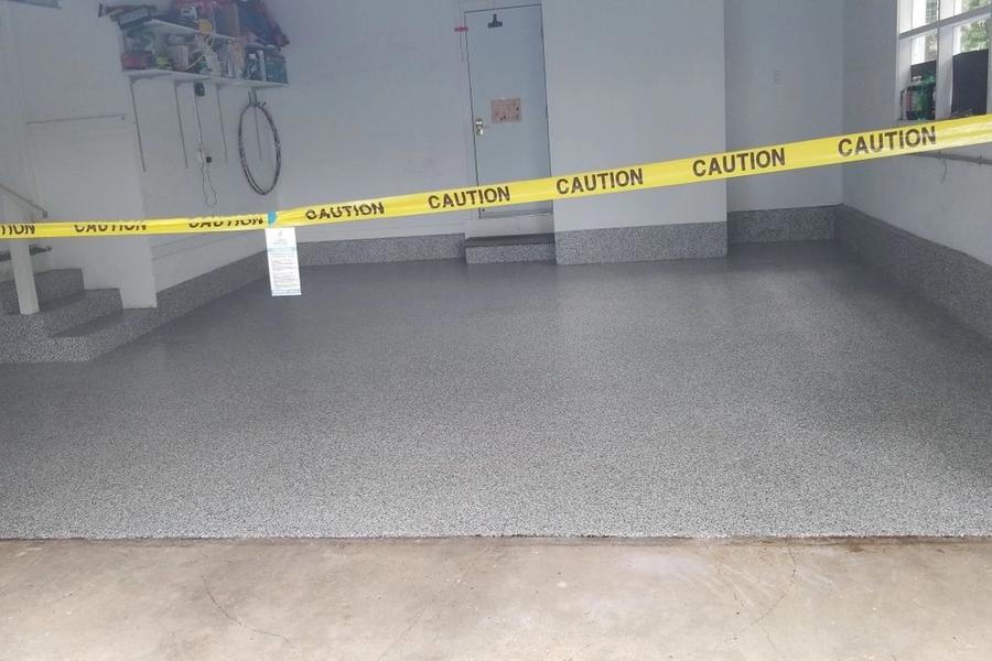 Freshly coated garage floor with a gray speckled flake finish, cordoned off by yellow tape reading "CAUTION" while the new coating cures.