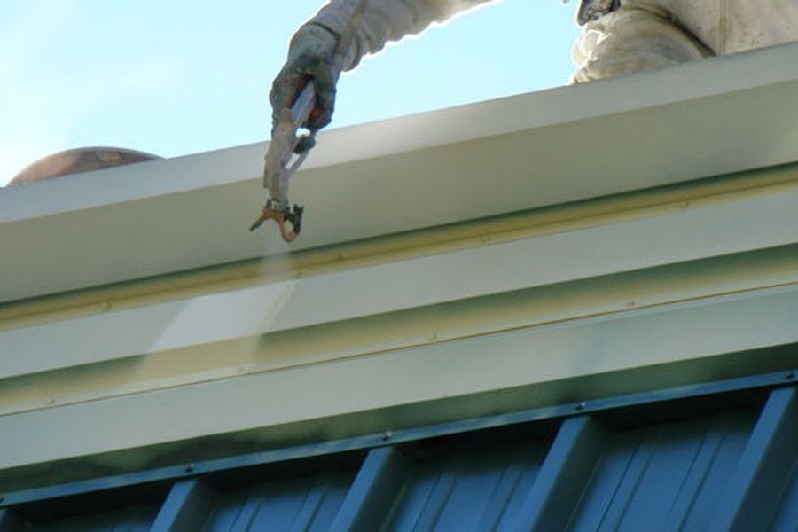 Painter's arm reaching down from a rooftop, spray-painting the cream-colored metal fascia above a blue standing-seam roof against a clear sky.