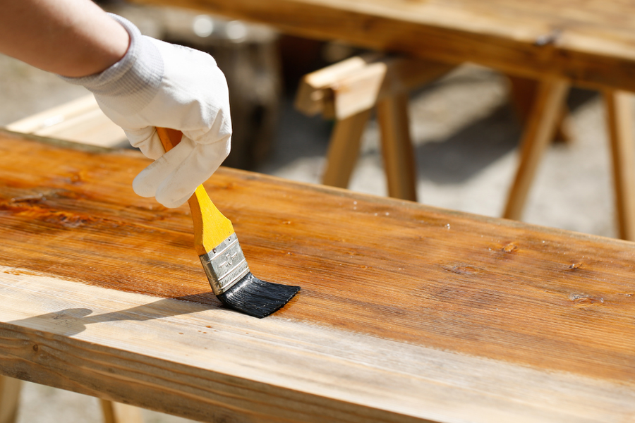 A gloved hand brushes dark wood stain onto a weathered plank outdoors, with the fresh finish contrasting against the unstained boards and sawhorses behind.