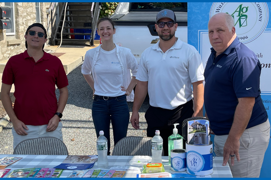 Four team members stand behind an outdoor fair table with brochures, paint cans, and water bottles, beside an A.G. Williams Painting Company banner dated 1906.