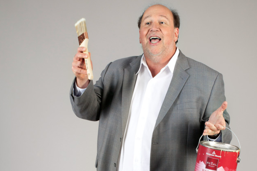 A smiling man in a gray blazer and white shirt holds up a paintbrush in one hand and a red Benjamin Moore Regal paint can in the other against a plain gray backdrop.