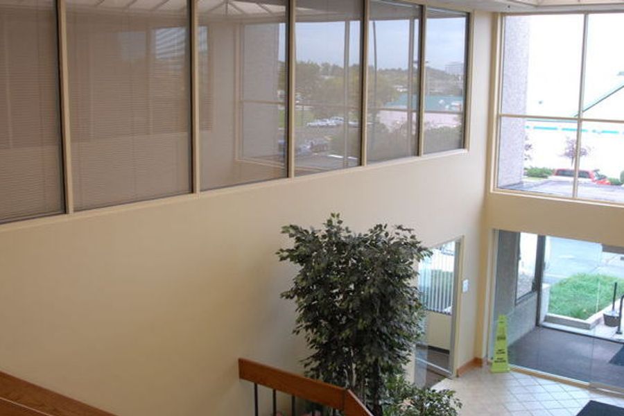 Two-story office lobby with cream walls, a bank of interior windows with blinds, potted trees, glass entry doors, and a lit exit sign.