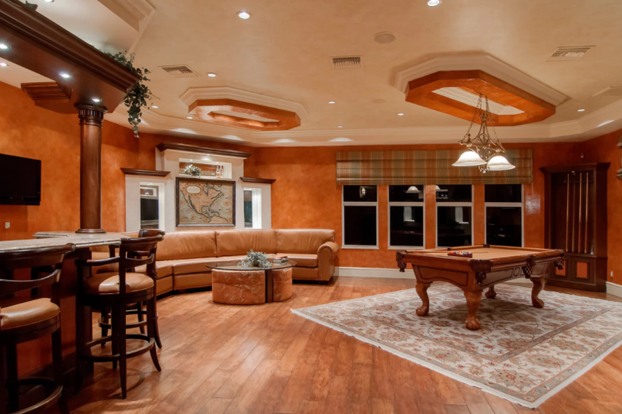 Spacious game room with warm terracotta faux-finished walls, a leather sectional sofa, pool table on a patterned rug, bar with stools, and coffered ceiling accents.
