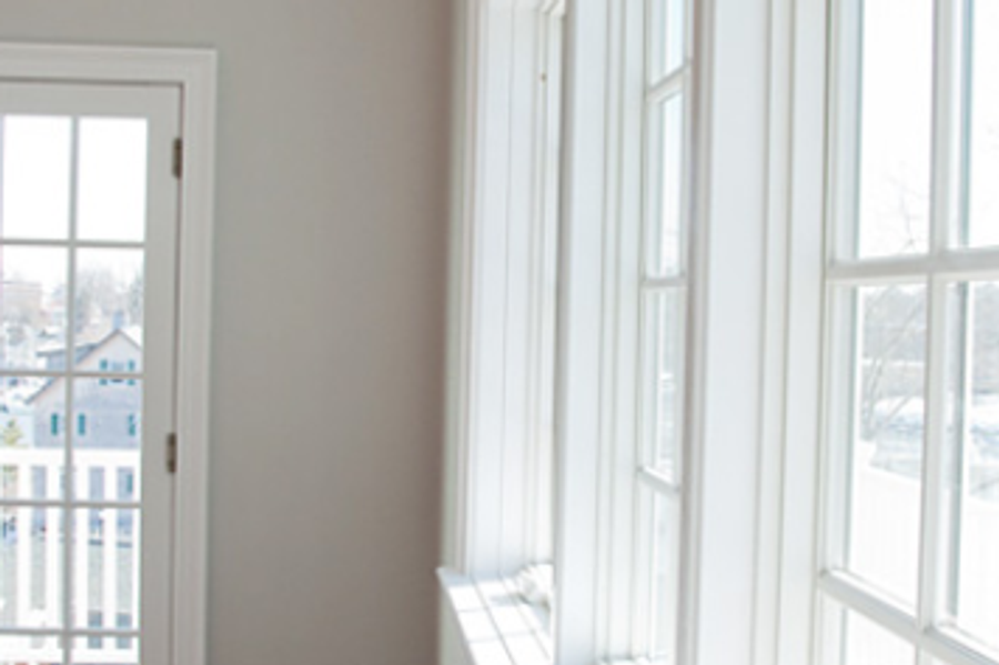 Sunlit corner of an empty room with light gray walls, a glass-paned door, and tall white-trimmed windows overlooking neighboring houses.