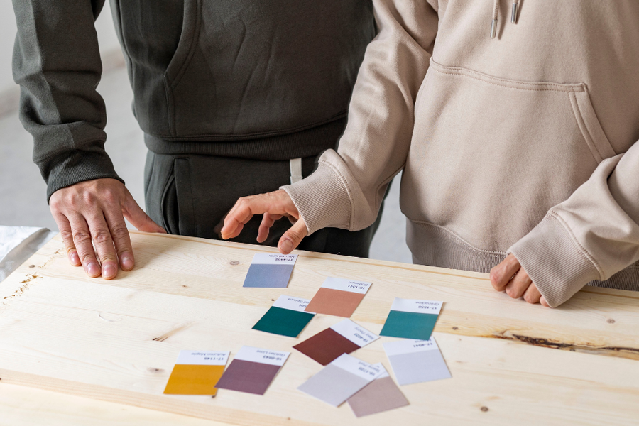 Two people in hooded sweatshirts stand at a light wood table, one pointing to an arrangement of labeled paint swatches in muted teal, plum, mustard, and neutral tones.