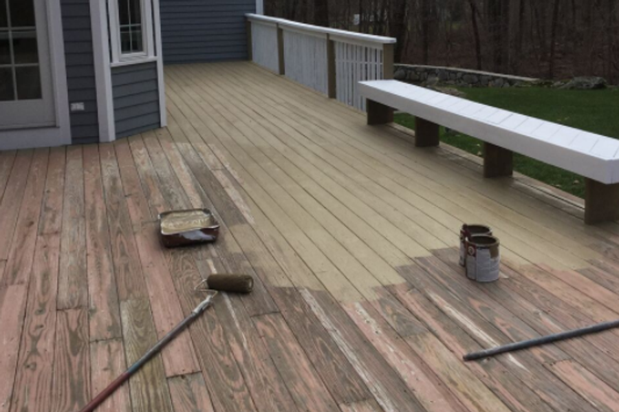 Backyard deck mid-project, with fresh tan stain covering most of the boards, weathered pink wood still exposed, and a roller, tray, and paint cans on the deck.