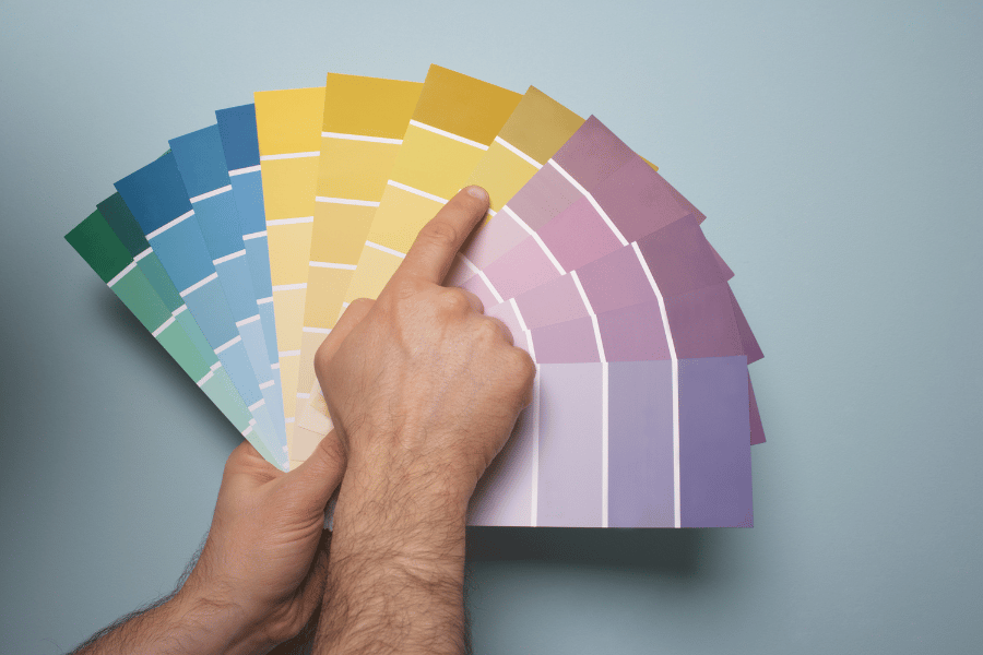 Two hands fanning out paint color swatches in greens, blues, yellows, and purples against a pale blue background, with one finger pointing to a yellow shade.