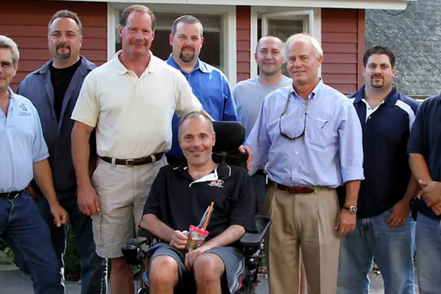 Nine men in casual and work shirts gather around a smiling man seated in a power wheelchair outside a red-sided house.
