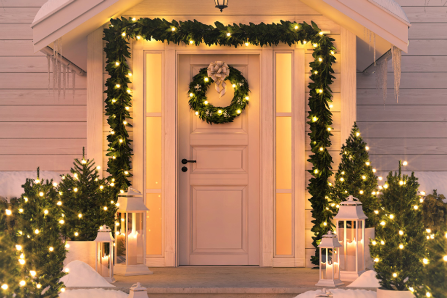 Front porch decorated for the holidays, with lit evergreen garland framing a cream-colored door, a wreath, small lighted trees, and white lanterns lining snowy steps.