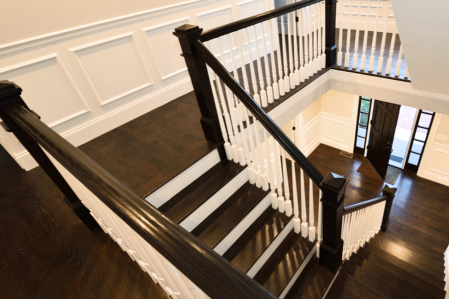 View down a staircase with dark-stained handrails and treads, white-painted spindles and risers, white wainscoting, and dark hardwood floors in a bright entryway.