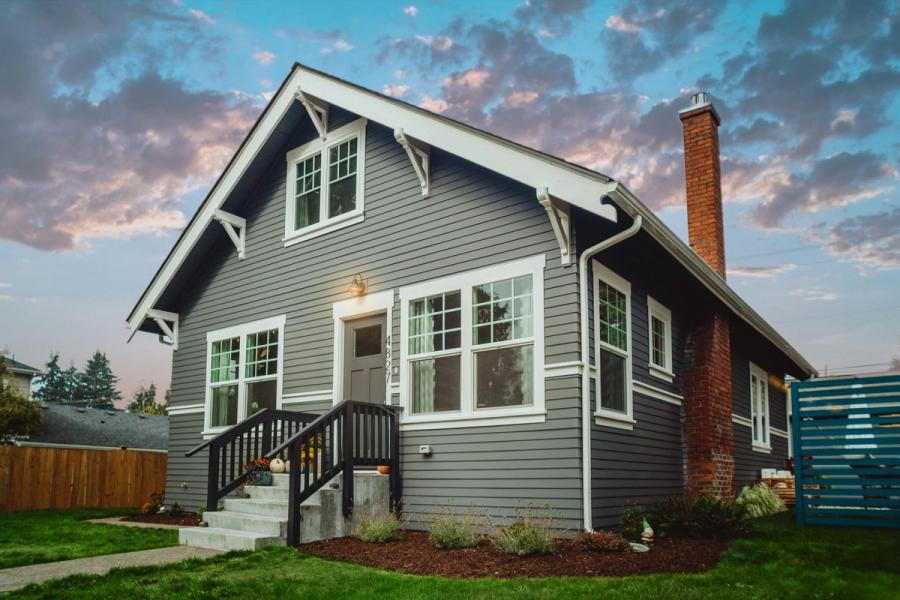 Gray craftsman-style house with white trim, a brick chimney, and a dark front porch railing, lit at dusk under a pink and blue sky.