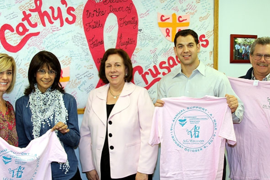 Five people hold up pink Support-A-Walk T-shirts bearing the A.G. Williams logo in front of a large "Cathy's Crusaders" banner covered in handwritten messages.