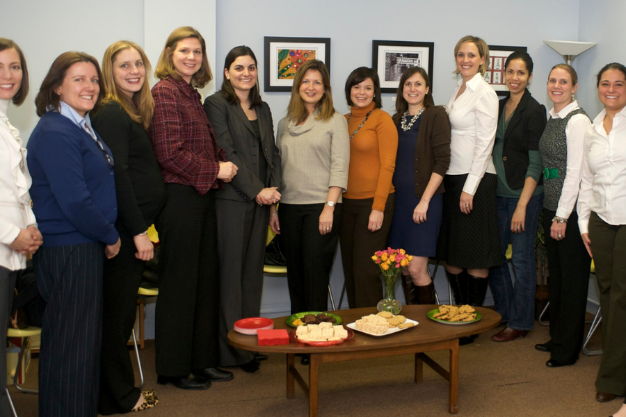 Fourteen women in business attire stand together smiling in a freshly painted light blue office with framed artwork, gathered behind a coffee table with flowers and refreshments.