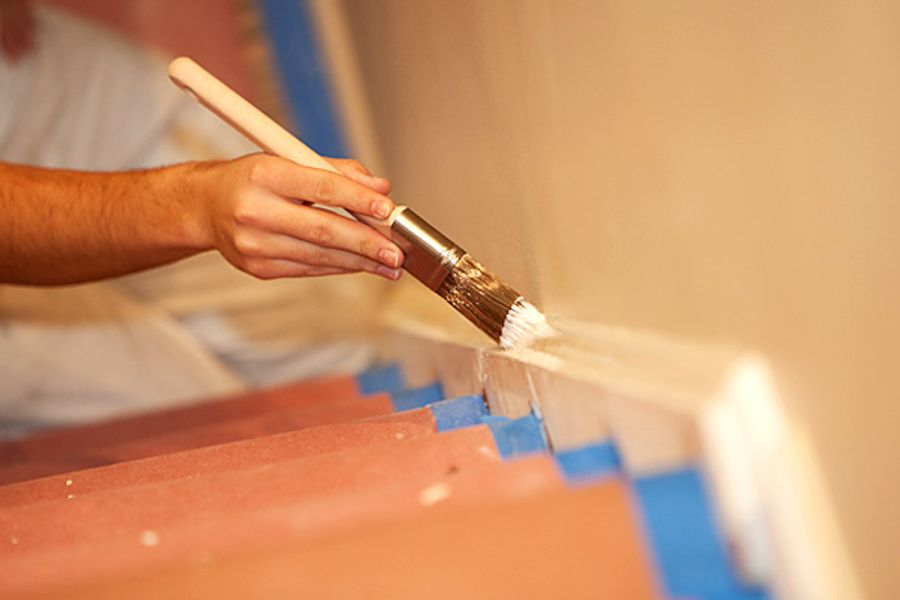 A painter's hand cutting in along stair trim with a round brush and white paint, with blue tape protecting the reddish stair treads.