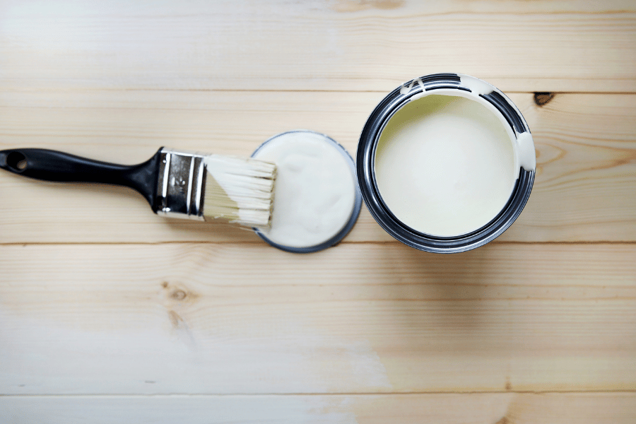 Overhead view of an open can of white paint and a black-handled brush resting on a small lid, set on light wood planks.