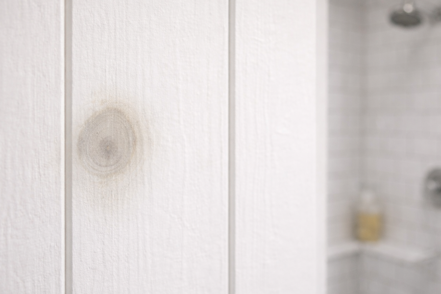 Close-up of a brown knot stain bleeding through white-painted wood paneling, with a tiled shower and chrome fixtures blurred in the background.