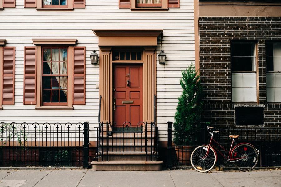 White clapboard house with a red front door, brown shutters, and black iron fence, with a red bicycle leaning against the fence beside a brick building.