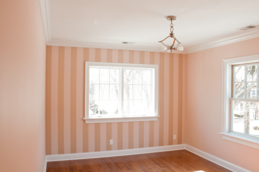 Empty freshly painted room with vertical peach-and-cream striped accent wall, white crown molding and trim, hardwood floors, and two windows overlooking a residential street.