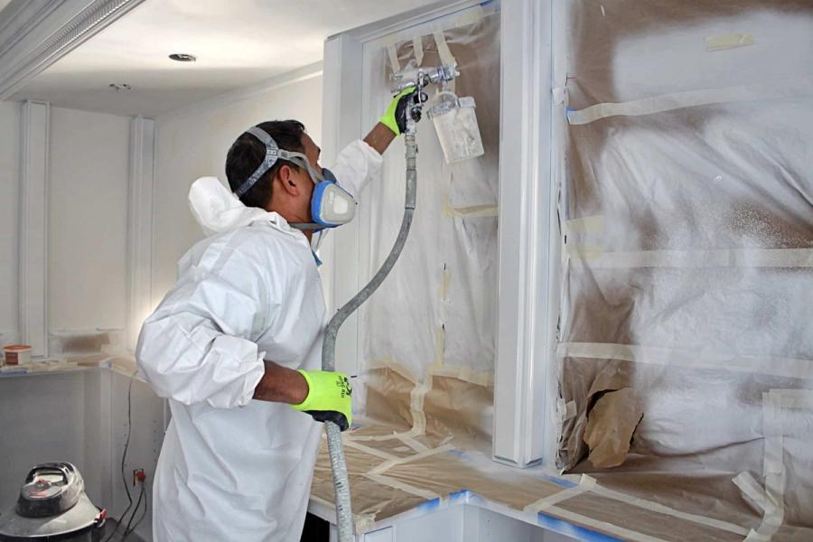 A painter in a white protective suit, respirator, and green gloves spray-paints interior cabinetry, with surrounding surfaces masked in paper and tape.