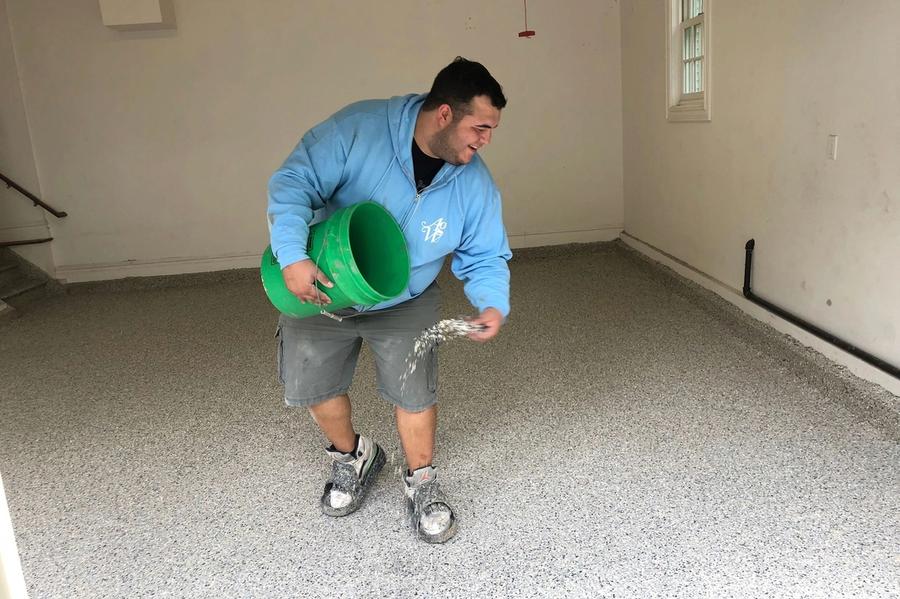 A worker in a blue hoodie scatters decorative flakes from a green bucket onto a freshly coated speckled garage floor.