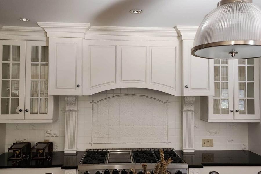 White painted kitchen cabinets with glass-front doors surround a stainless range and arched range hood, with black countertops and a ribbed glass pendant light.