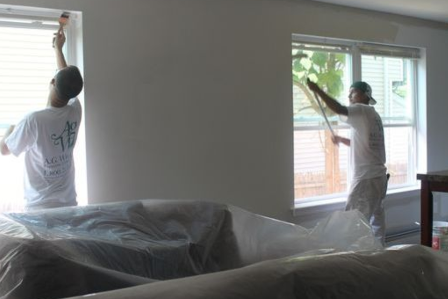 Two painters in white A.G. Williams T-shirts brush the trim around living room windows, with the furniture covered in protective plastic sheeting.