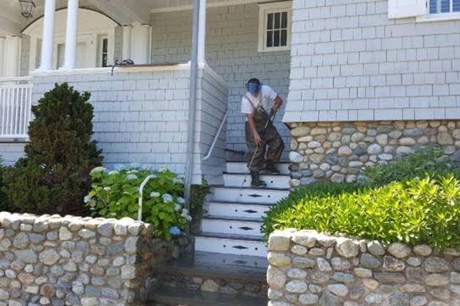 A worker in waders and a face shield power washes white front steps of a gray shingled house bordered by stone walls and hydrangeas.