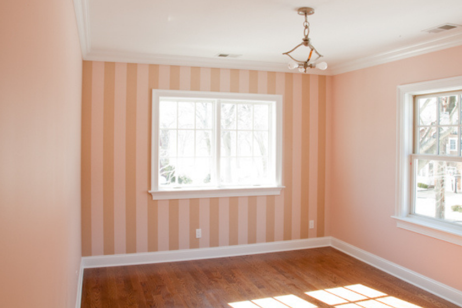 Empty sunlit room with one wall in soft pink and cream vertical stripes, adjoining solid pink walls, white crown molding and trim, and hardwood floors.