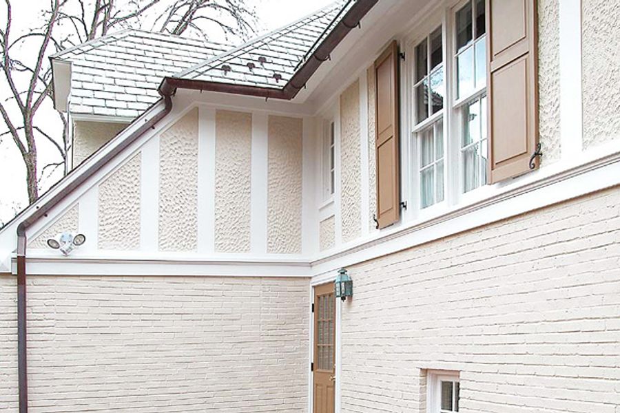 Corner of a home with cream-painted brick on the first story, textured stucco with white half-timber trim above, tan shutters, and a slate roof.