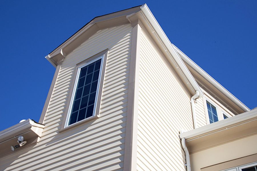 Upward view of a home's cream-colored clapboard siding, white trim, gutters, and gridded windows against a clear blue sky.