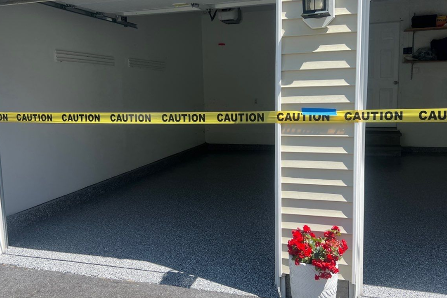 Yellow caution tape stretched across an open two-bay garage with a freshly coated blue-gray flake floor, cream siding, and potted red flowers by the entrance.
