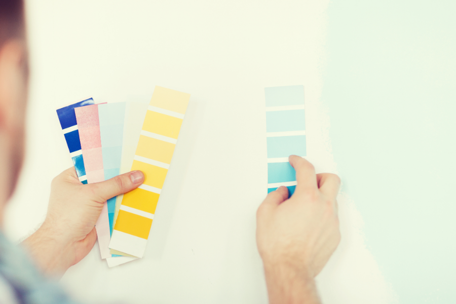 A person compares fanned paint swatch cards in yellow, blue, and pink tones against a white wall, holding a blue swatch strip up to a pale painted section.