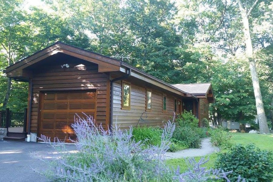 Ranch-style home with warm wood-stained siding and a matching garage door, surrounded by tall trees, lavender plants, and a stone walkway on a sunny day.
