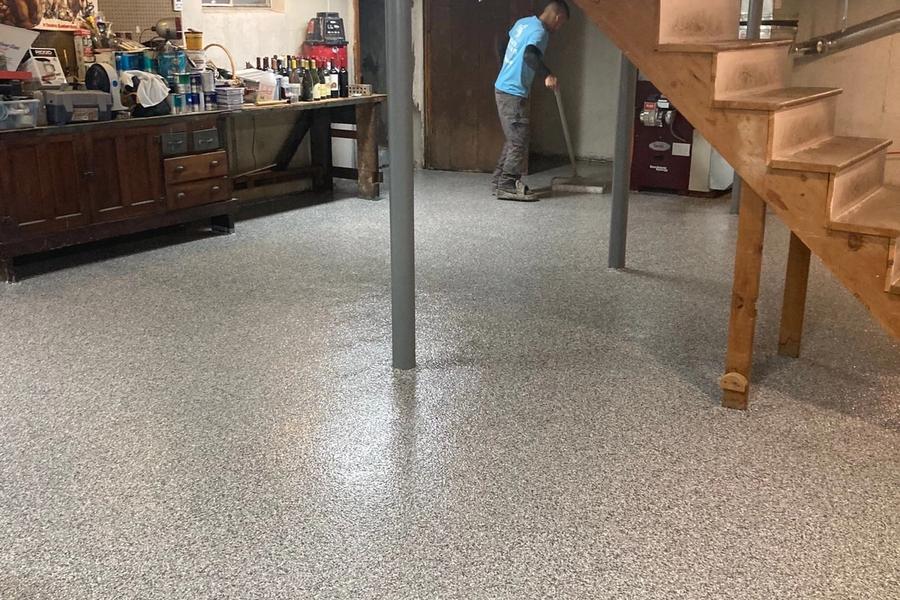 Worker in a blue shirt applying a gray flake coating to a basement floor near a wooden staircase, workbench, and exposed ceiling joists.