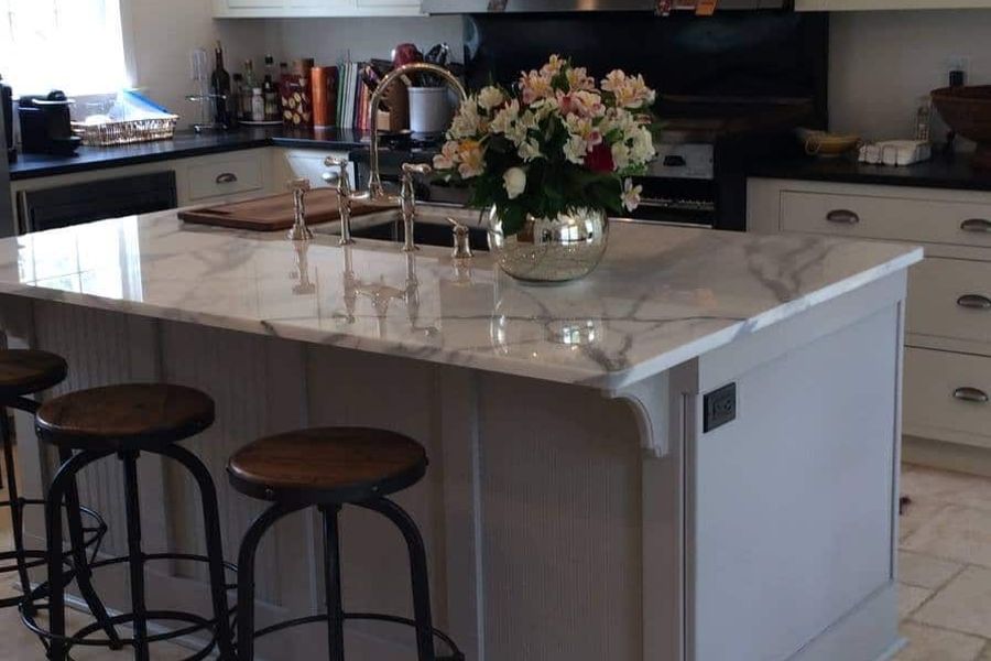 Kitchen with a gray island topped in white marble, wood-seat metal barstools, pendant lights, cream cabinets, and a vase of fresh flowers beside the sink.